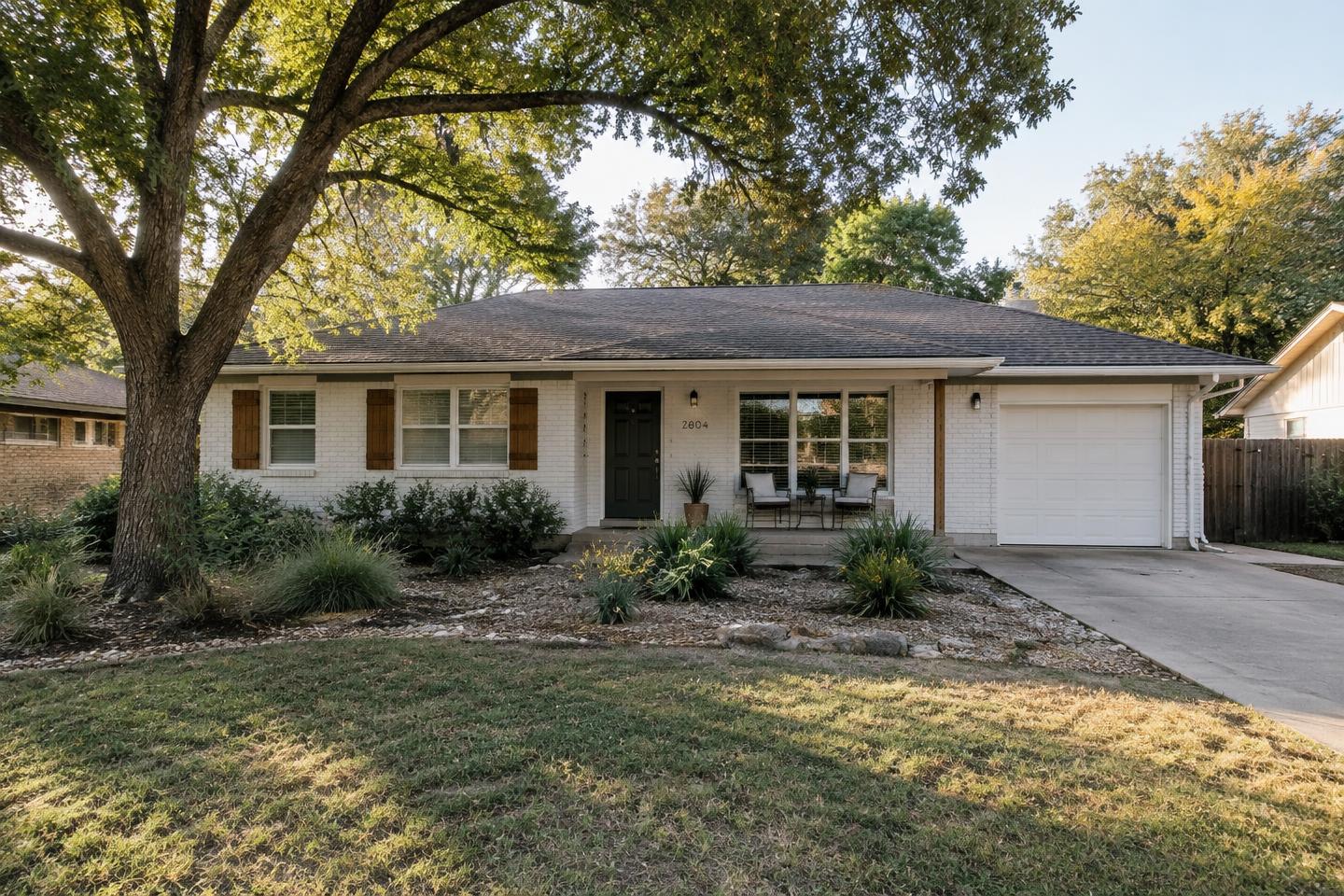 Modest single-story white brick ranch home beneath a mature pecan tree in Zilker